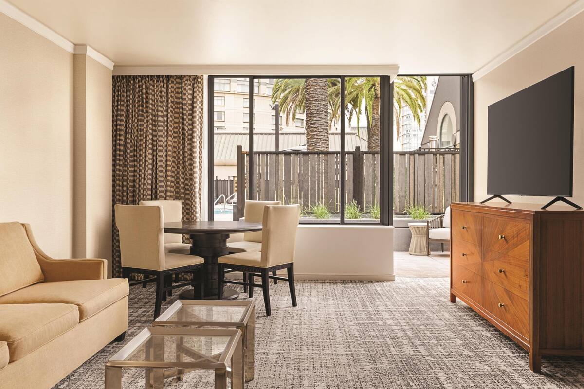 A hotel room with a sofa, a dining table with chairs, a TV, a wooden cabinet, and a large window with a view of palm trees and buildings.