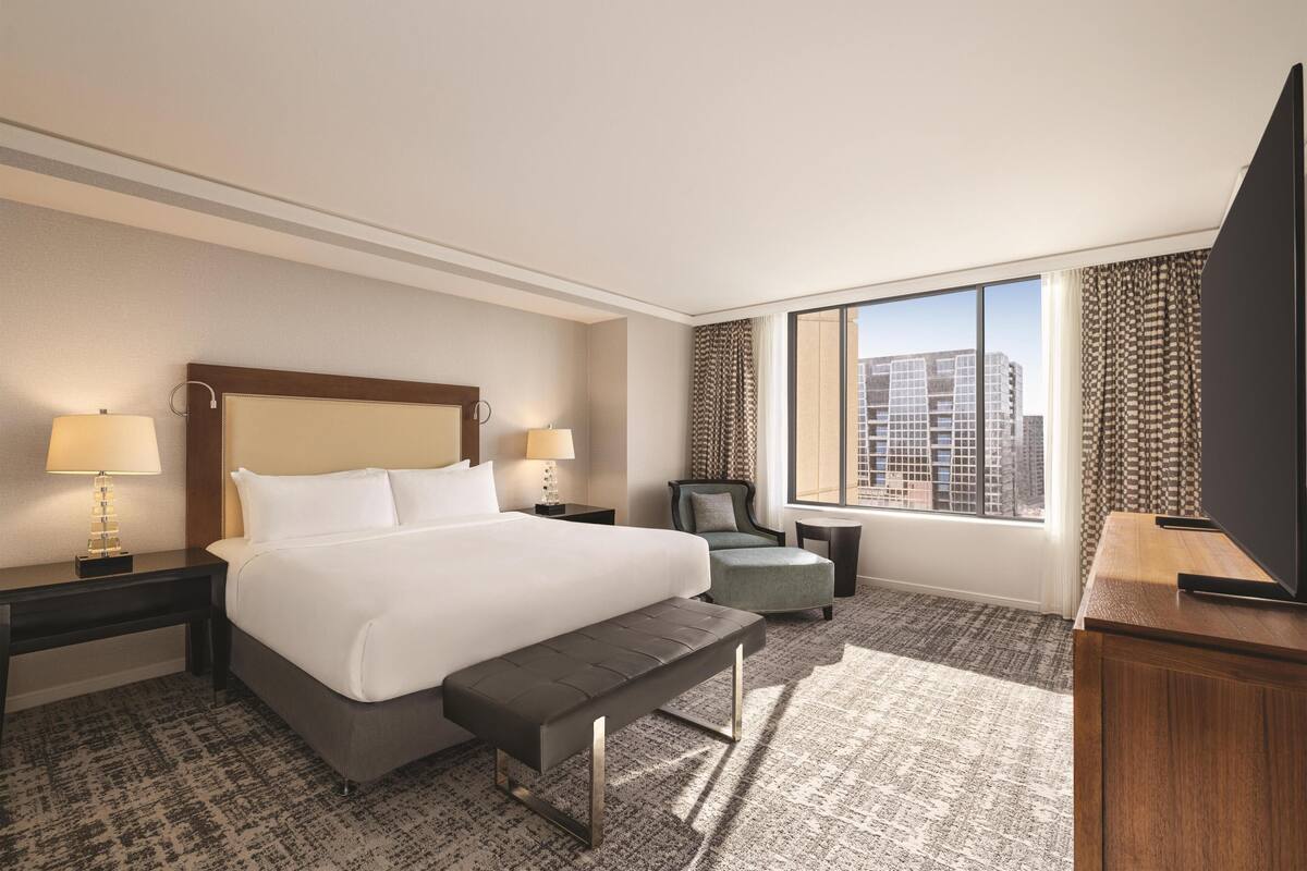 A hotel room with a large bed, a bench, a chair, a desk, a TV, and a window with a city view.