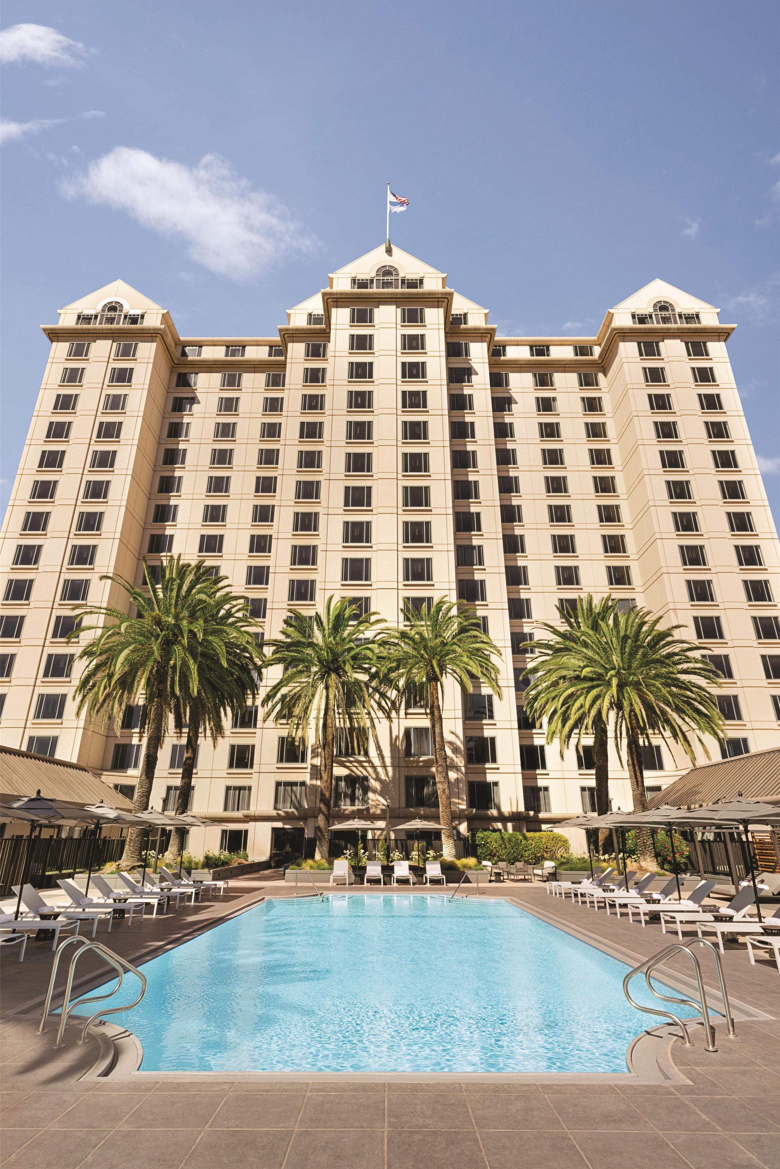 A large hotel with a swimming pool, palm trees, and a flag on top.