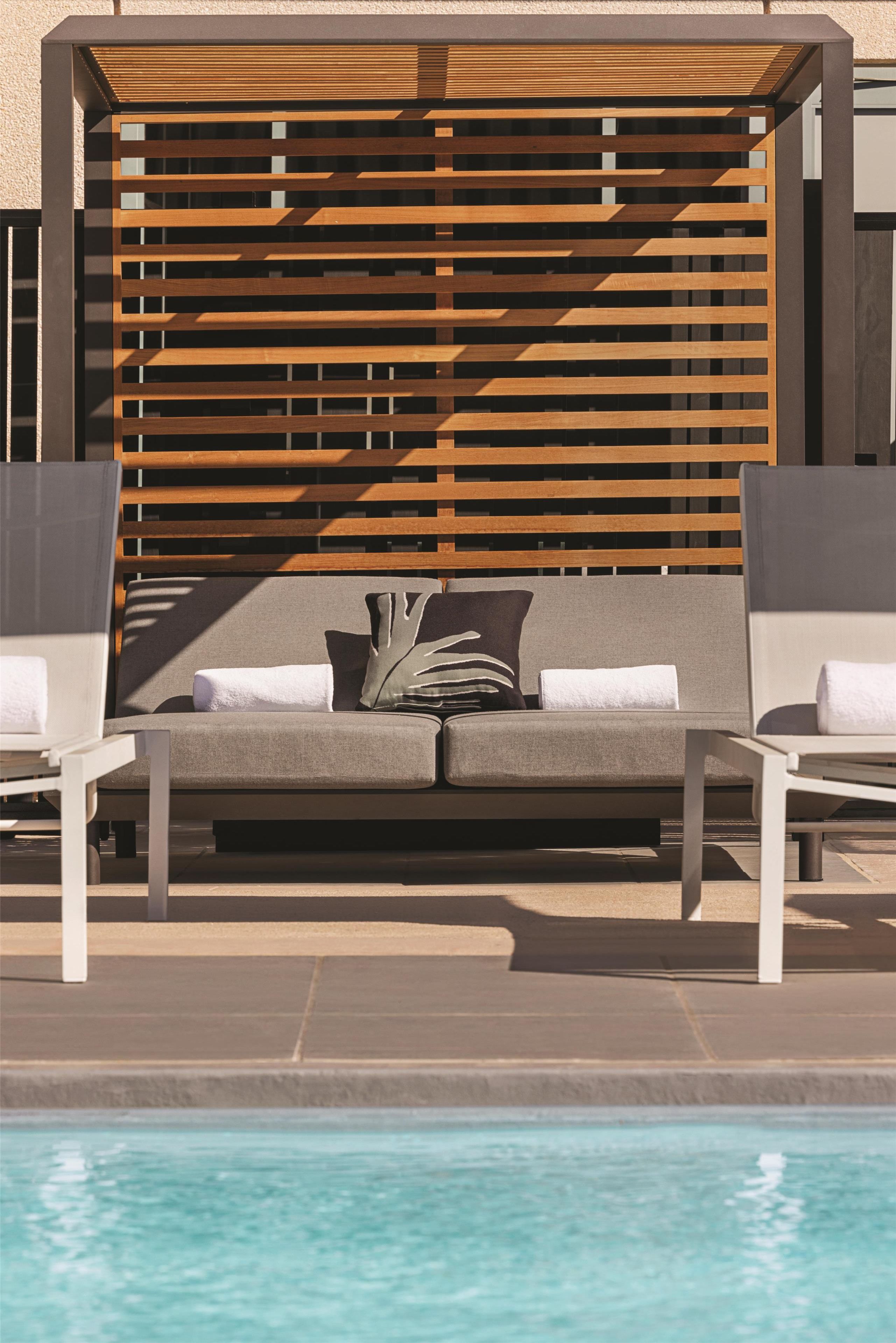 A poolside lounge area with a sofa, two chairs, and a wooden pergola structure.