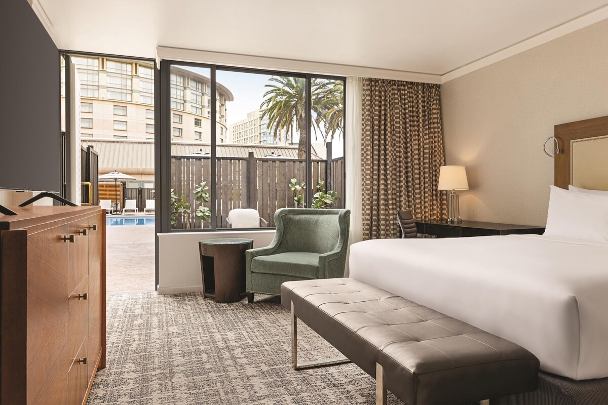 A hotel room with a large bed, a green armchair, a small table, and a view of a pool and palm trees.