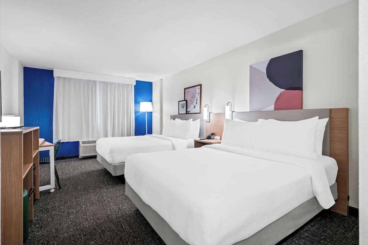 A hotel room with two beds, a desk, a chair, a lamp, and artwork on the wall.