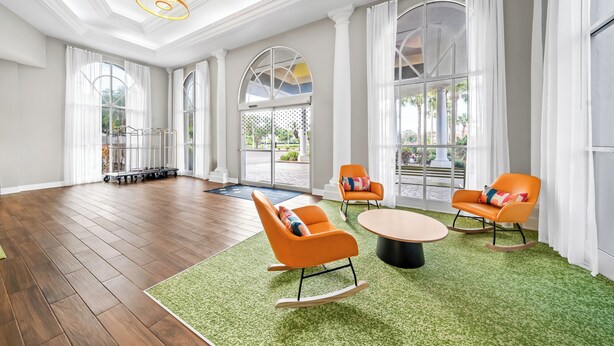 Une pièce spacieuse avec un plancher en bois, de grandes fenêtres et une moquette verte. On y trouve deux fauteuils à bascule orange et une table ronde.