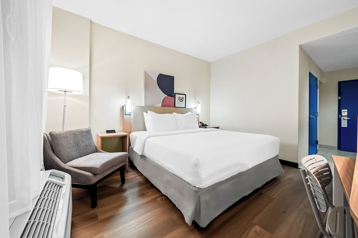 A hotel room with a bed, a chair, a desk, a lamp, and a wall-mounted artwork.