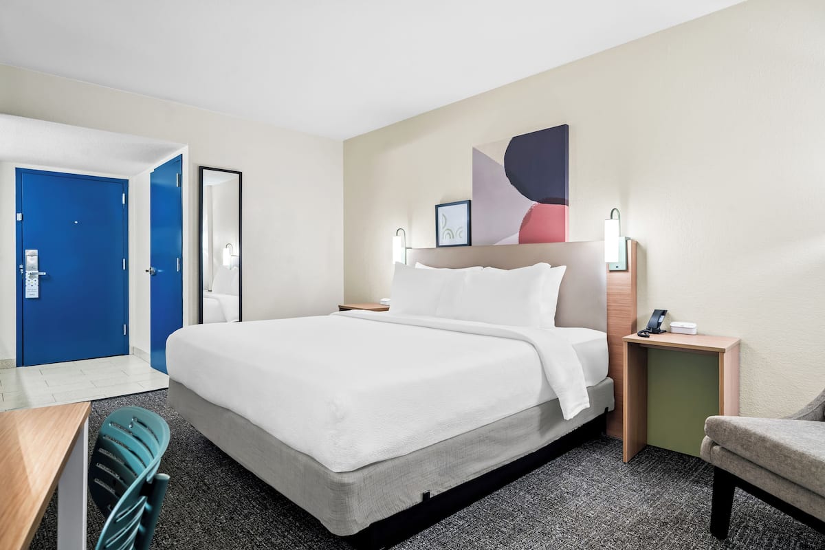 A hotel room with a large bed, a desk, a chair, and a nightstand with a lamp.