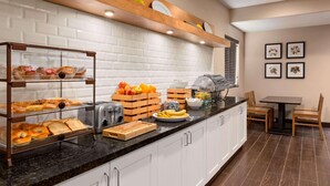 A breakfast bar with pastries, fruits, and a toaster. There is a wooden shelf above with plates and a window with blinds.