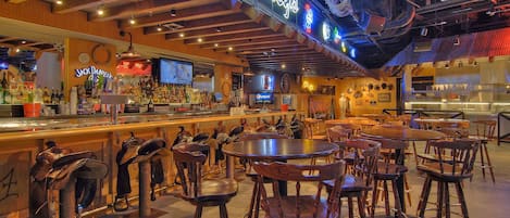 Una zona de bar con sillas y mesas de madera, un letrero de neón y diversas botellas y vasos en estantes.