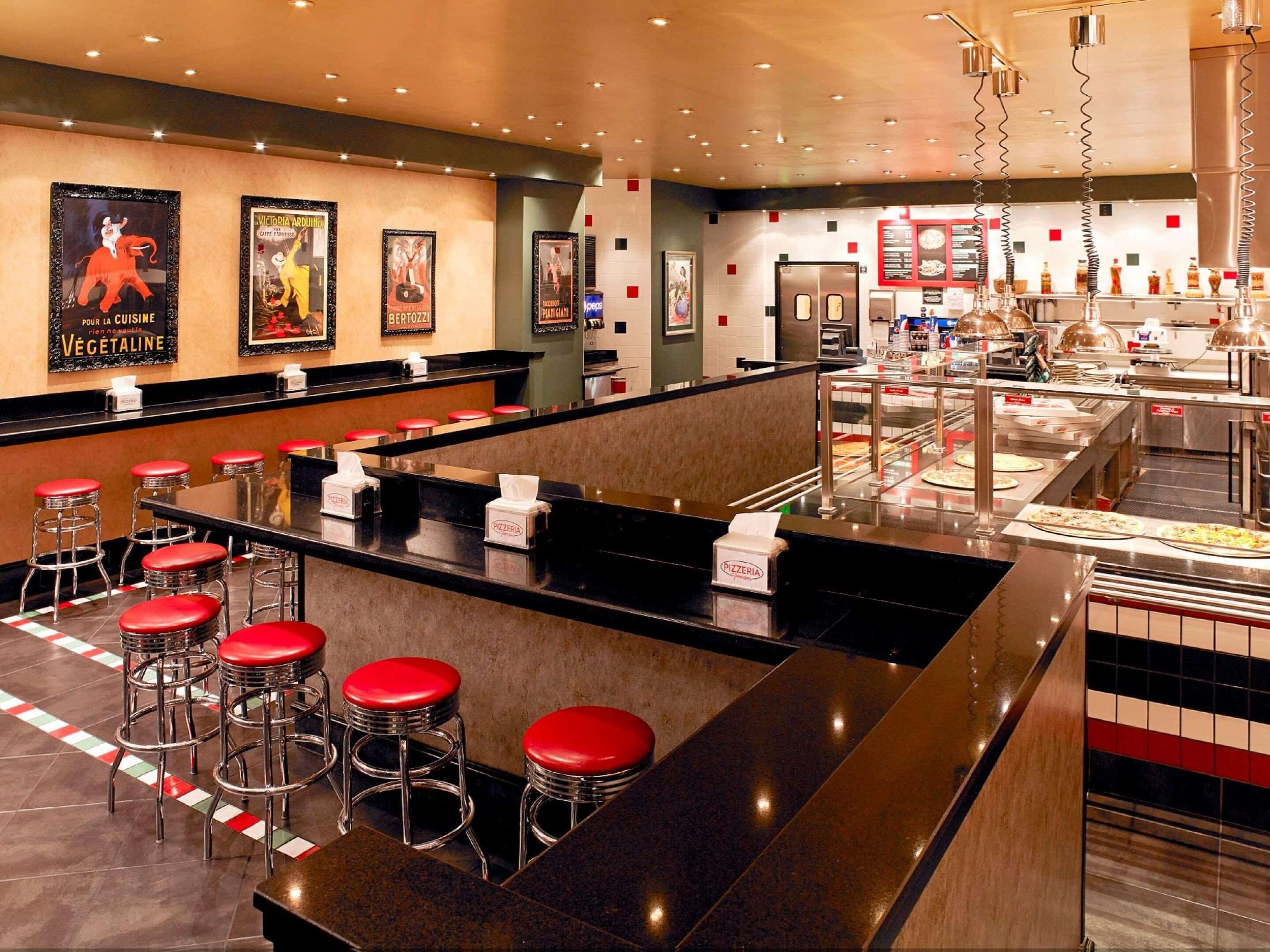 A modern pizzeria with a counter, red stools, and pizza display.