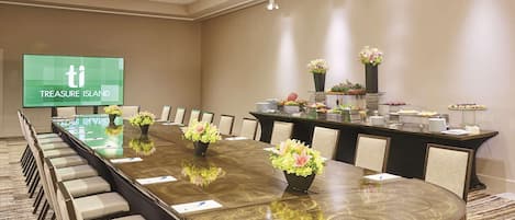 A conference room with a long table, chairs, a screen displaying 'TREASURE ISLAND', and a buffet area with flowers and food.