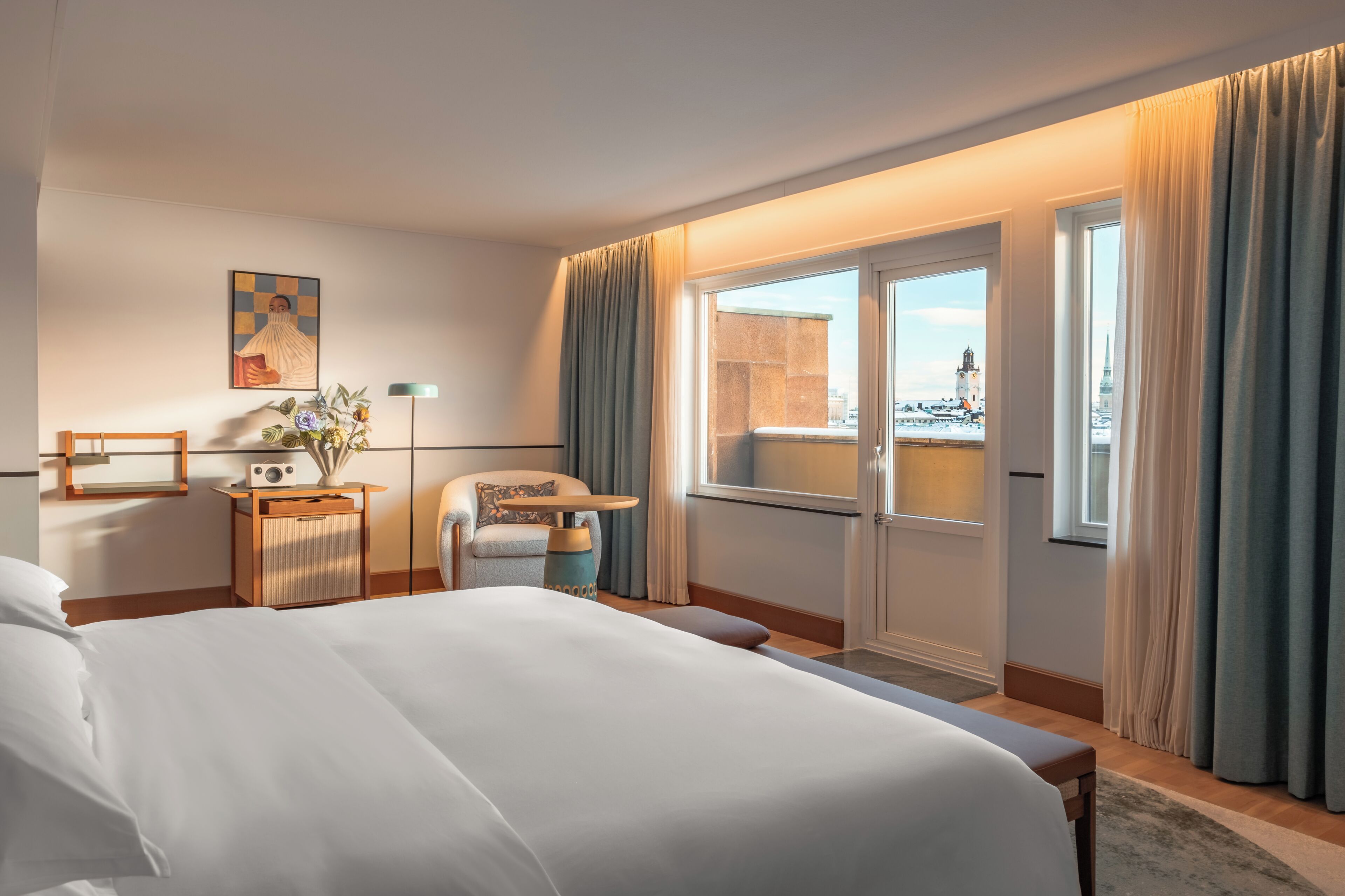 A hotel room with a large bed, a desk with a lamp, a chair, a window with curtains, and a view of a cityscape.