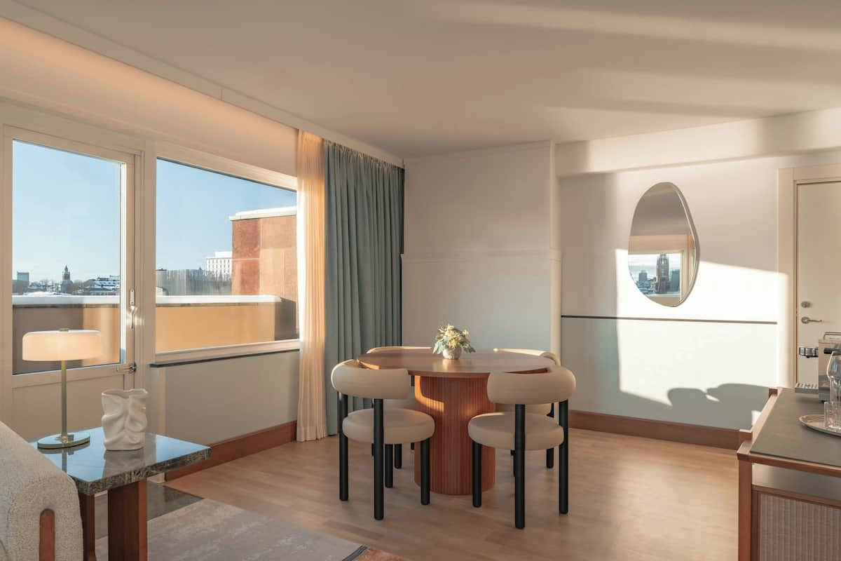 A modern hotel room with a round dining table, four chairs, a small side table, a large window with curtains, and a view of the cityscape.