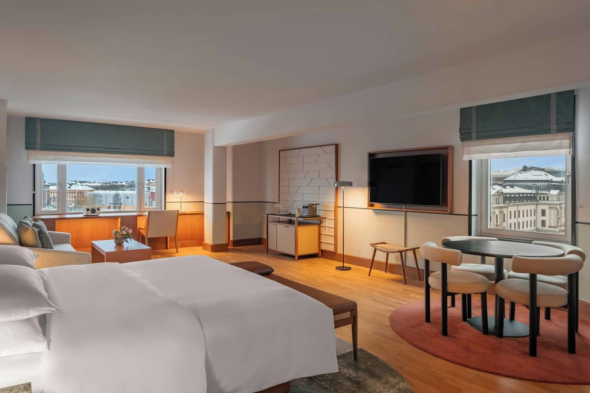 A modern hotel room with a large bed, a dining area, a kitchenette, and a TV.