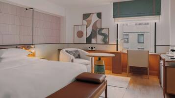 A hotel room with a bed, a desk, a chair, and a sofa. There are wall art pieces and a window with a view of buildings.