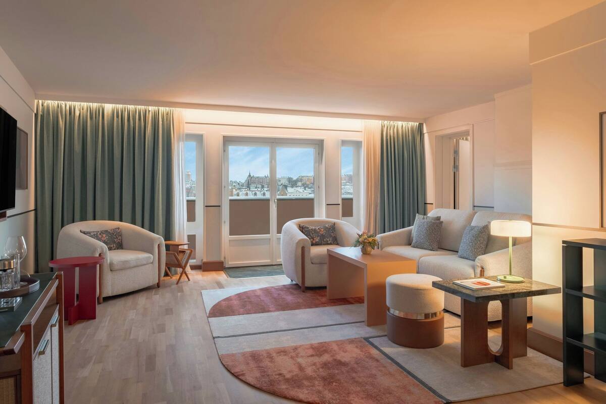 A modern living room with two sofas, a coffee table, and a TV. There is a large window with curtains, and a view of buildings outside.