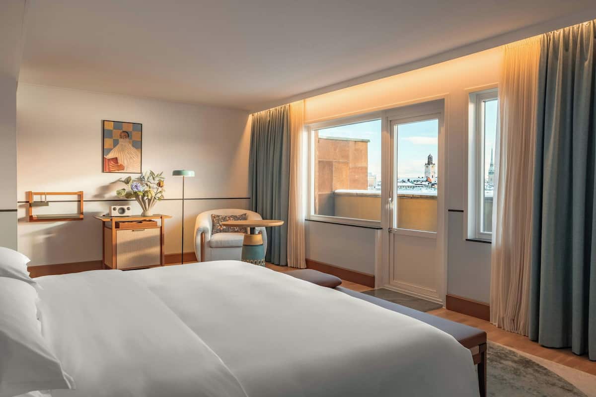 A hotel room with a large bed, a desk with a lamp, a chair, a window with curtains, and a view of a cityscape.