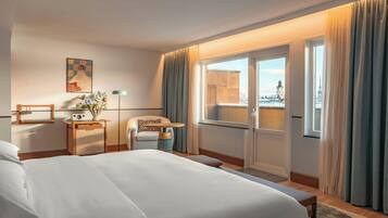 A hotel room with a large bed, a desk with a lamp, a chair, a window with curtains, and a view of a cityscape.
