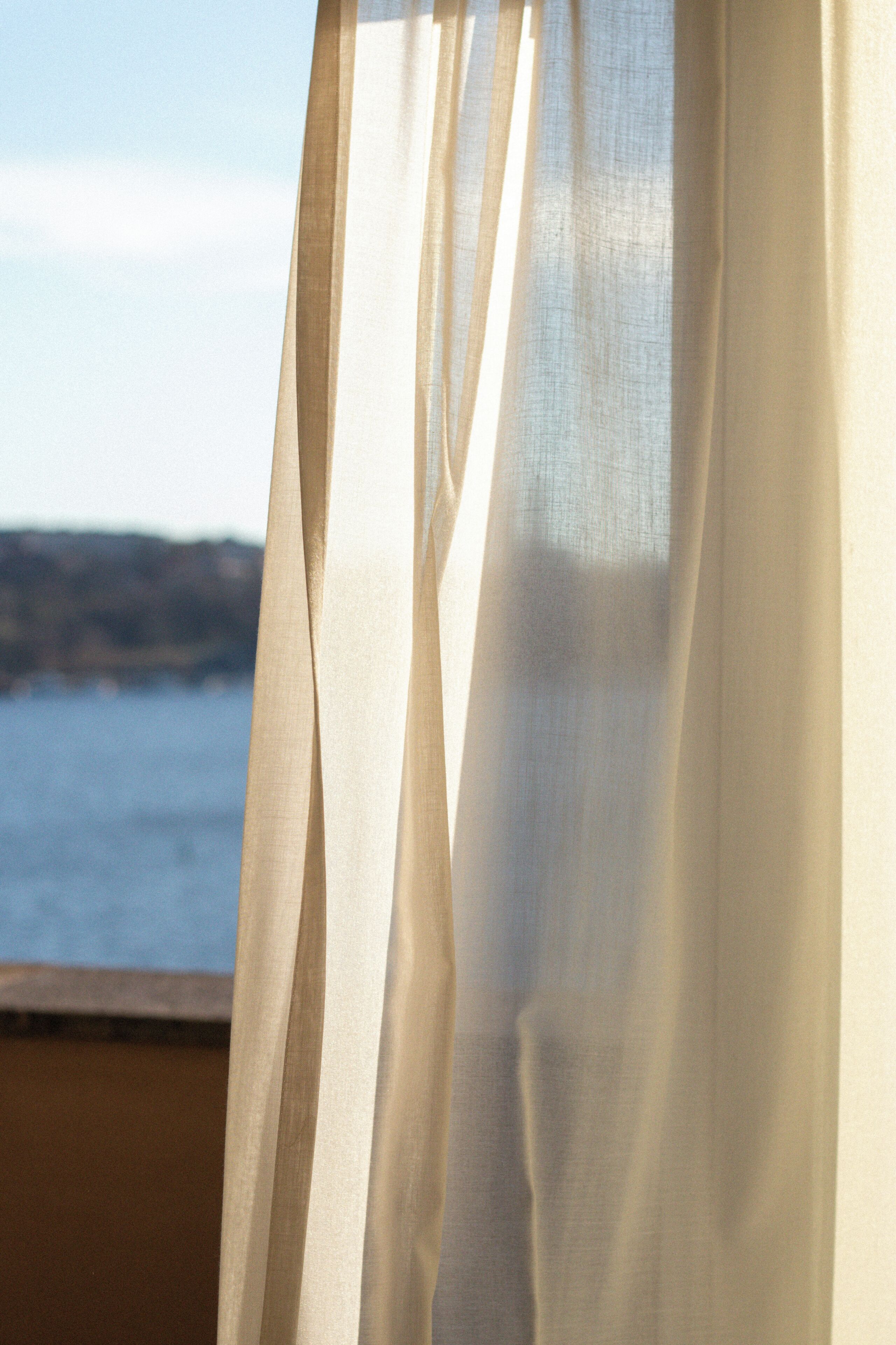 A sheer curtain partially drawn, revealing a view of a body of water and distant land.