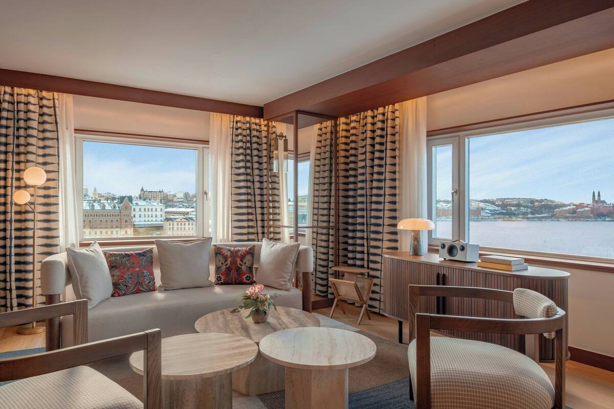 A modern hotel room with a large window offering a city view, a cozy seating area with a sofa and chairs, and a small table with a lamp.