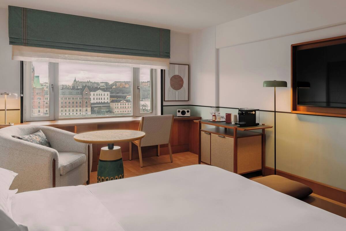 A hotel room with a bed, a seating area with a table and chairs, a kitchenette, and a large window offering a city view.