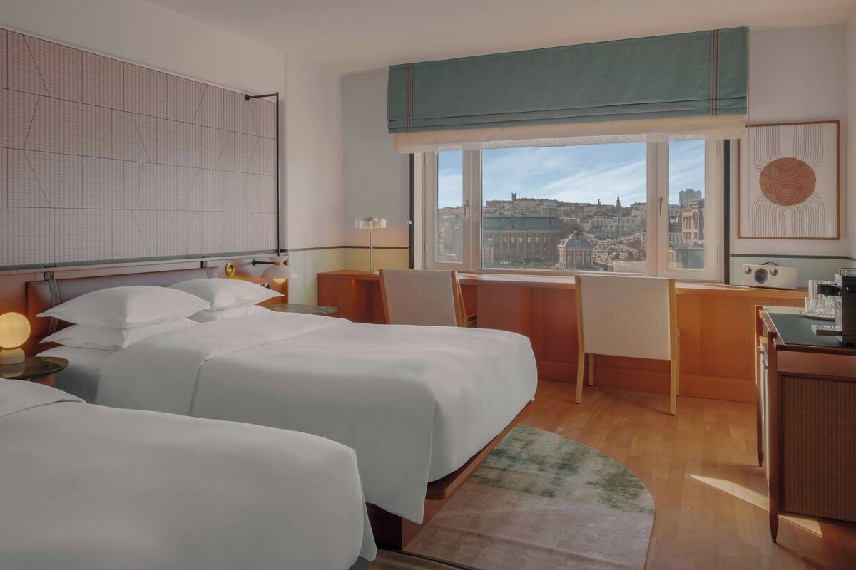 A hotel room with two beds, a desk, a chair, a window with a city view, and a large abstract painting.