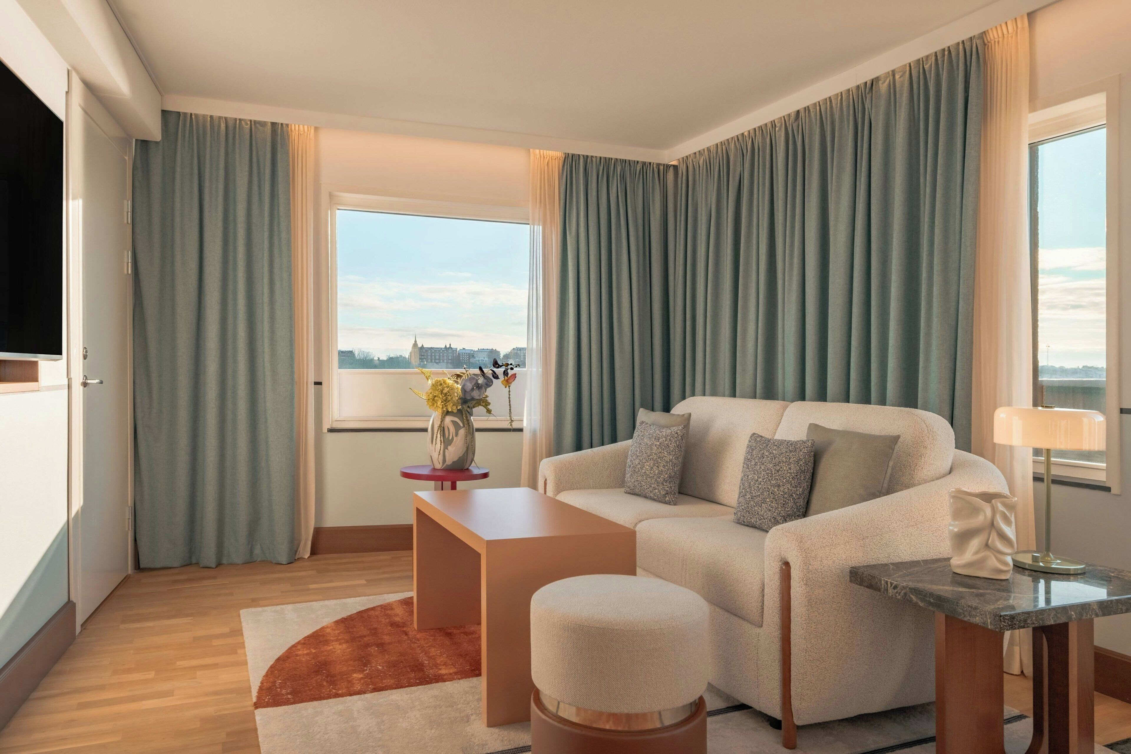 A modern living room with a sofa, ottoman, side table, and a view of the city through large windows.