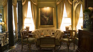 A room with a patterned carpet, a sofa, two chairs, a table with a fruit bowl and wine glasses, and a painting on the wall.