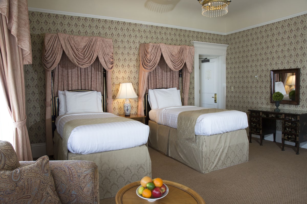A hotel room with two beds, a desk, a chair, and a fruit bowl on a small table.