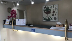 Una recepción de hotel con un cartel de "NO FUMAR", un reloj, una tarjeta de agradecimiento enmarcada, una planta en maceta y una ilustración botánica en la pared.