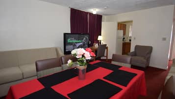 Habitación de hotel con mesa de comedor para dos, un sofá, un televisor y una cocina visible a través de una puerta abierta.