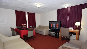 Habitación de hotel con sala de estar que incluye un sofá, una mesa de comedor con sillas, un televisor y un escritorio con lámpara.