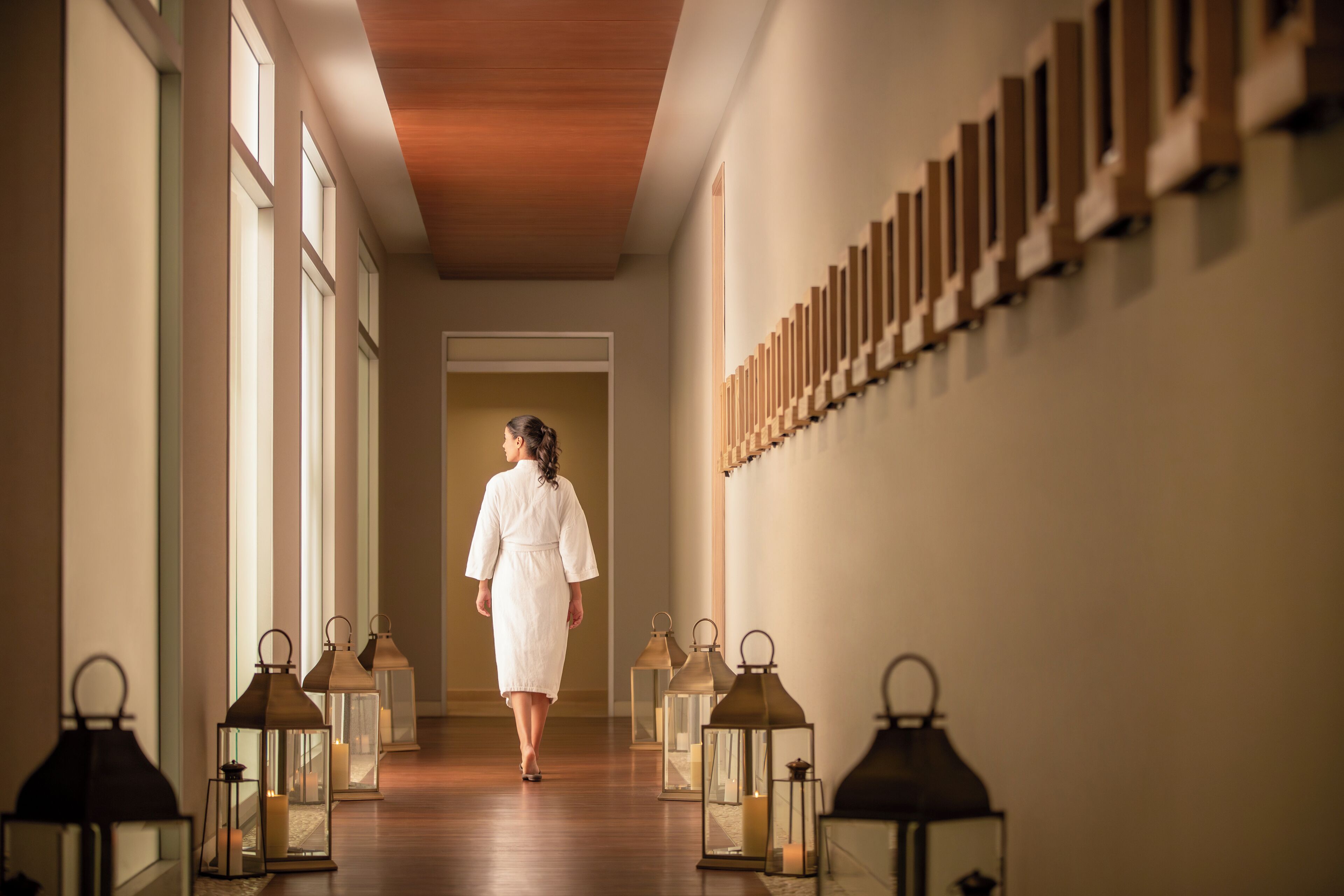 A corridor with a woman walking towards a door, flanked by lanterns and wooden panels.