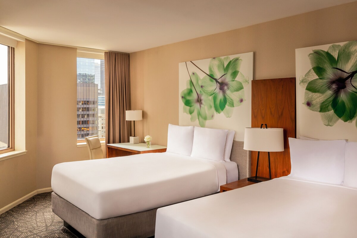 A hotel room with two beds, a large window with a view of buildings, a desk with a lamp, and artwork on the walls.