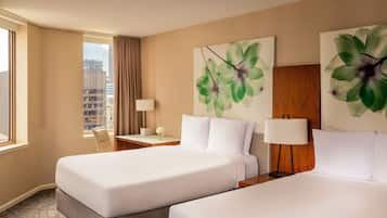 A hotel room with two beds, a large window with a view of buildings, a desk with a lamp, and artwork on the walls.