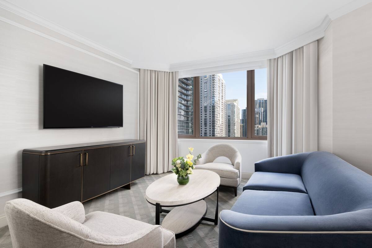 Una habitación de hotel moderna con un televisor de pantalla plana montado en la pared, un mueble de madera oscura, una mesa redonda con un jarrón de flores, un sofá azul y vistas de edificios altos a través de amplias ventanas.