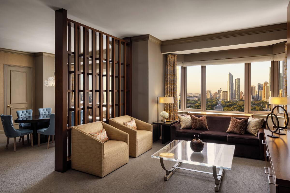Una habitación de hotel moderna con vistas a la ciudad, una zona de estar con dos sofás y una mesa de centro, y un comedor con mesa y sillas.