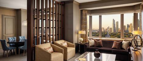 Una habitación de hotel moderna con vistas a la ciudad, una zona de estar con dos sofás y una mesa de centro, y un comedor con mesa y sillas.