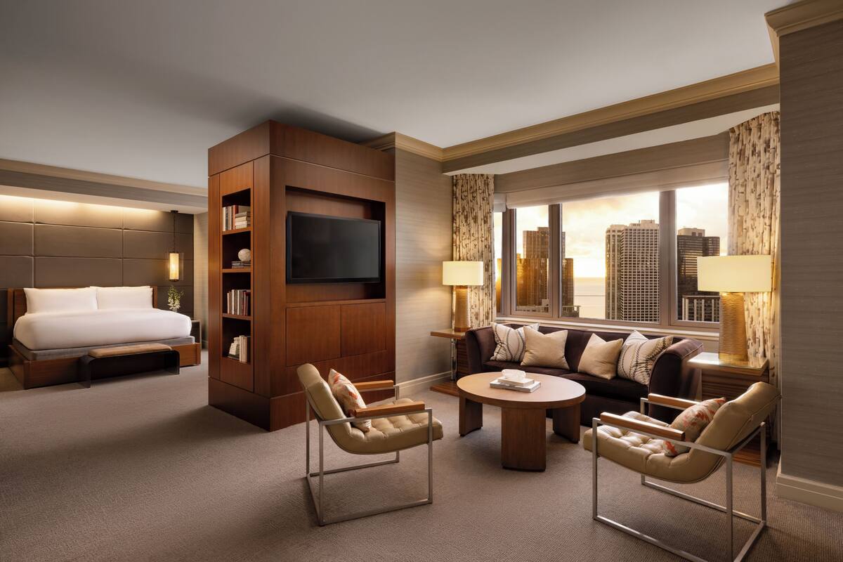 Una habitación de hotel con cama, televisión, sofá, dos sillones, mesa de centro y vista a la ciudad.