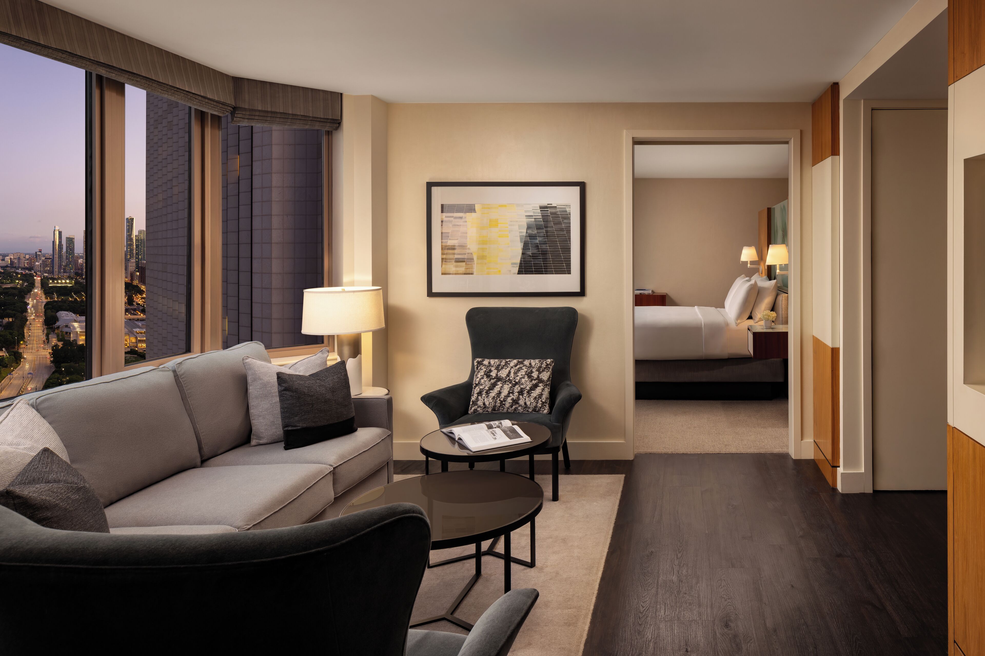 A hotel room with a sofa, armchair, and a small table. There is a bed in the background, a framed picture on the wall, and a view of the city through large windows.