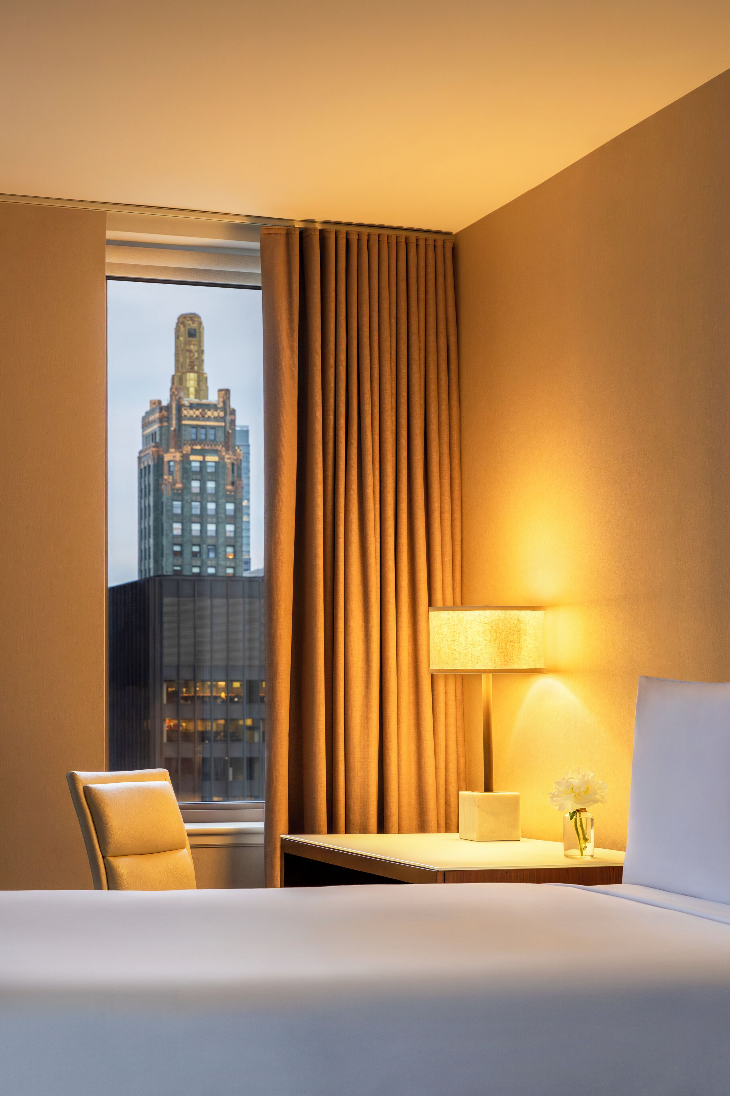 Une chambre d’hôtel avec un lit, une chaise, une table de chevet avec une lampe et une vue sur un immeuble par la fenêtre.
