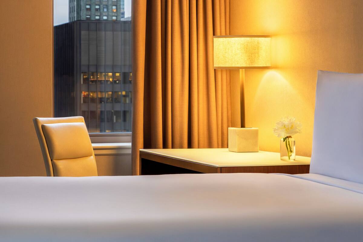 Habitación de hotel con cama, una silla, mesita de noche con lámpara y vista a un edificio por la ventana.