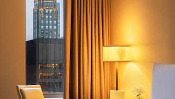 Et hotelværelse med en seng, en stol, et natbord med lampe, og udsigt til en bygning gennem et vindue.