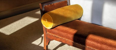 A wooden bench with a yellow cushion, positioned in a room with a white bed in the background.