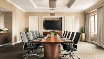 A conference room with a large wooden table, black leather chairs, a flat-screen TV, and a potted plant.