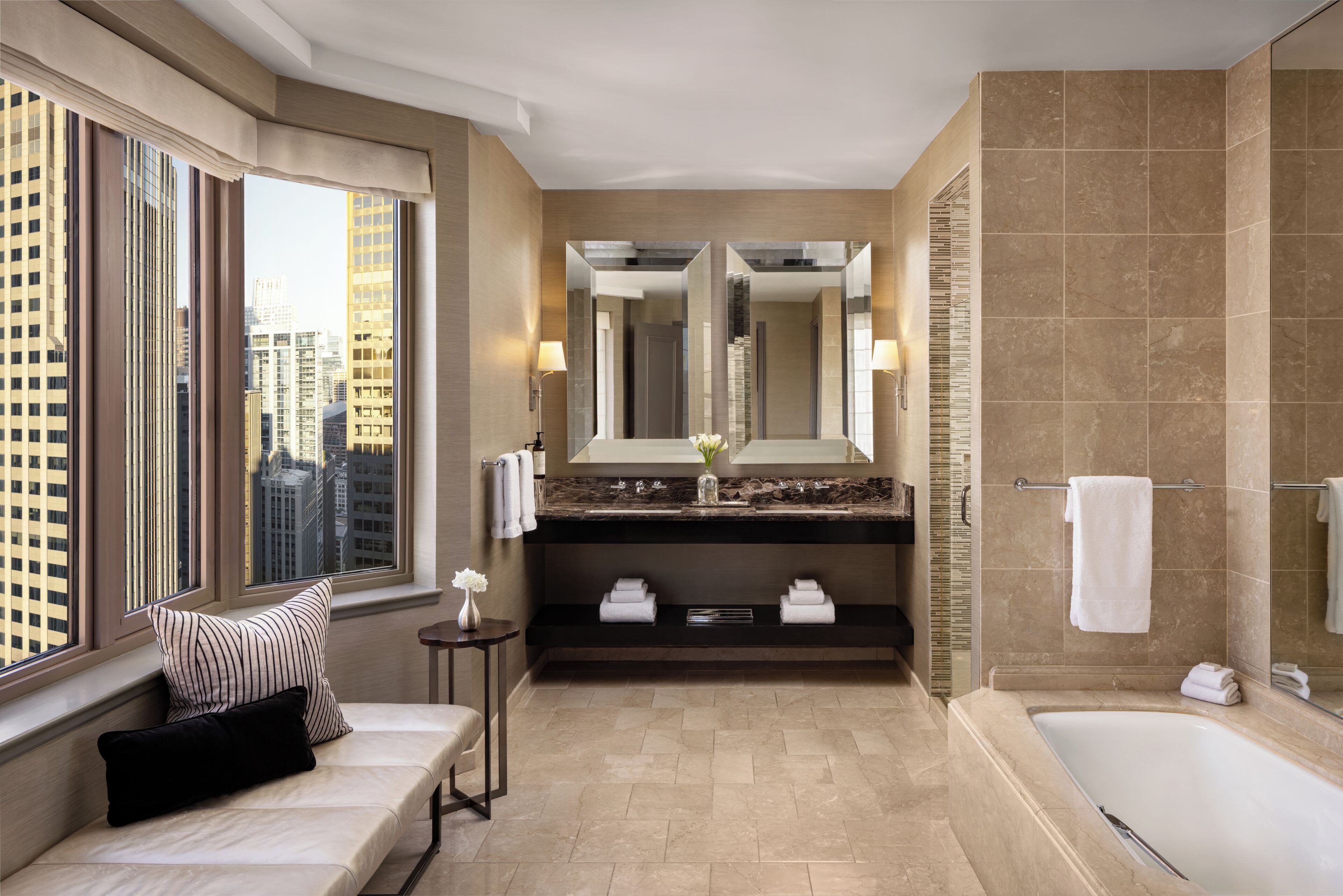 A modern bathroom with a large bathtub, a double vanity with a mirror, and a bench by the window.