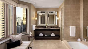 A modern bathroom with a large bathtub, a double vanity with a mirror, and a bench by the window.