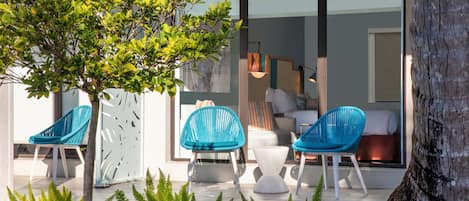 Een moderne buitenruimte met stoelen en een boom, een glazen deur naar een kamer met een bed en een hanglamp.