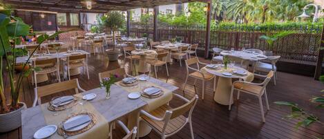 Un espacio al aire libre para comer con varias mesas y sillas, rodeado de follaje y una terraza de madera.
