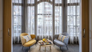 Una habitación con dos sillas, una mesa redonda con cosas para el desayuno y ventanas grandes con cortinas traslúcidas.