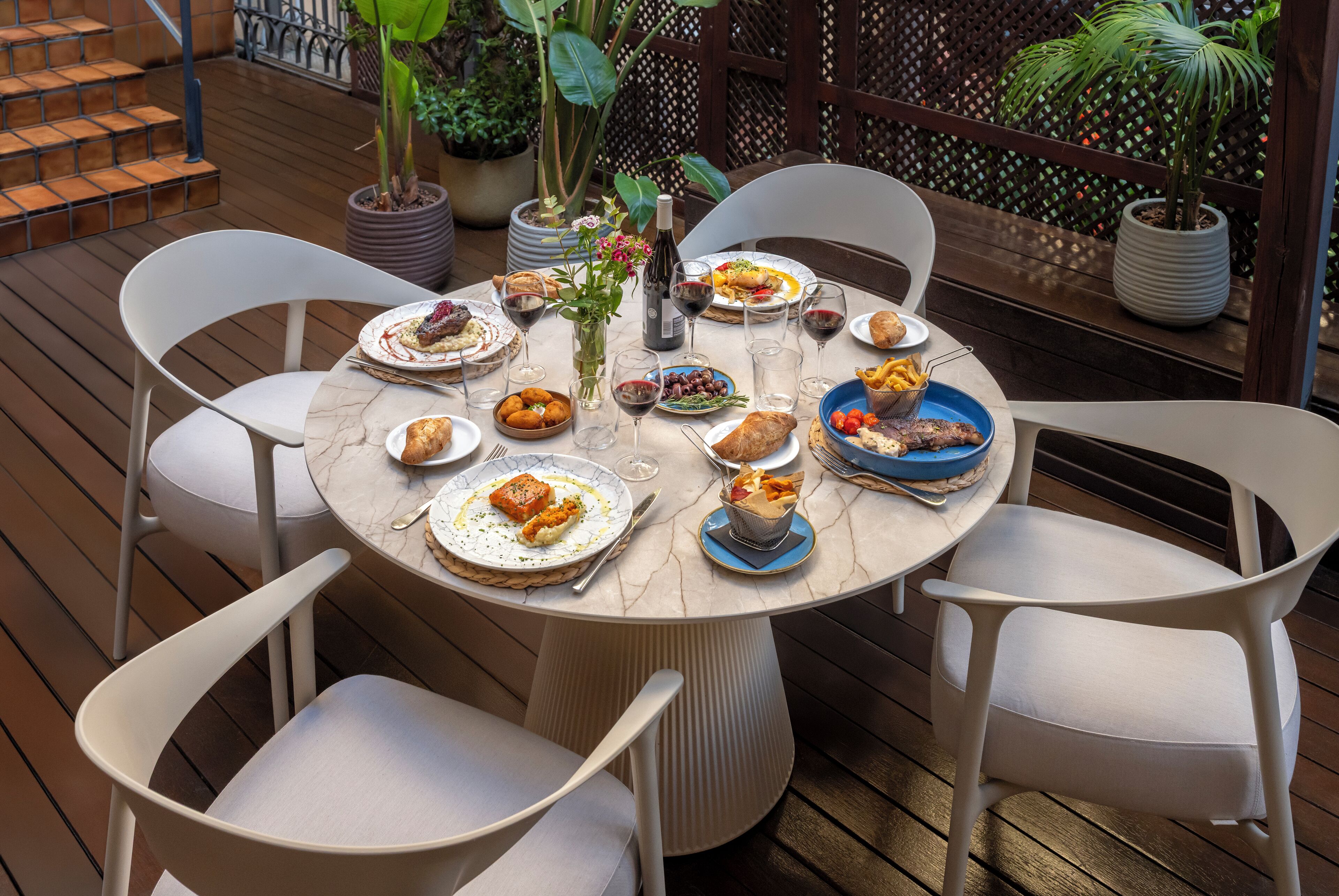 Een ronde tafel met witte stoelen, omgeven door potplanten en een houten terras.