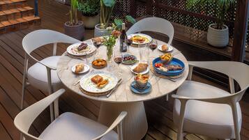 Una mesa redonda con sillas blancas, rodeada de plantas en macetas y una terraza de madera.